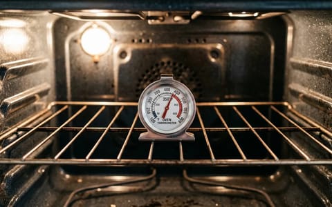 A stainless steel oven thermometer sitting on a wire rack inside a brightly lit oven.