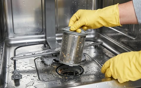 Person wearing yellow gloves removing the primary filter from the bottom of a dishwasher.