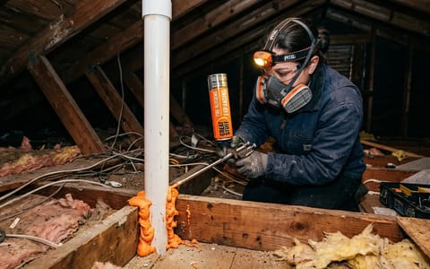 Homeowner wearing safety gear applying expanding foam around an attic plumbing pipe.