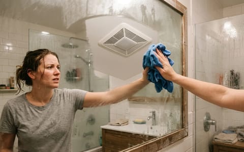 Homeowner wiping a fogged bathroom mirror with a ceiling exhaust fan in the background.