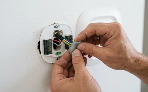 A homeowner sealing the wire hole behind a thermostat with plumber's putty to block wall cavity drafts.