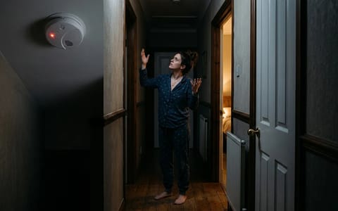 Homeowner looking up at a hardwired smoke detector in a dark hallway