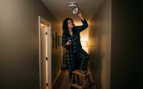 Homeowner replacing a smoke detector battery on a step stool at night